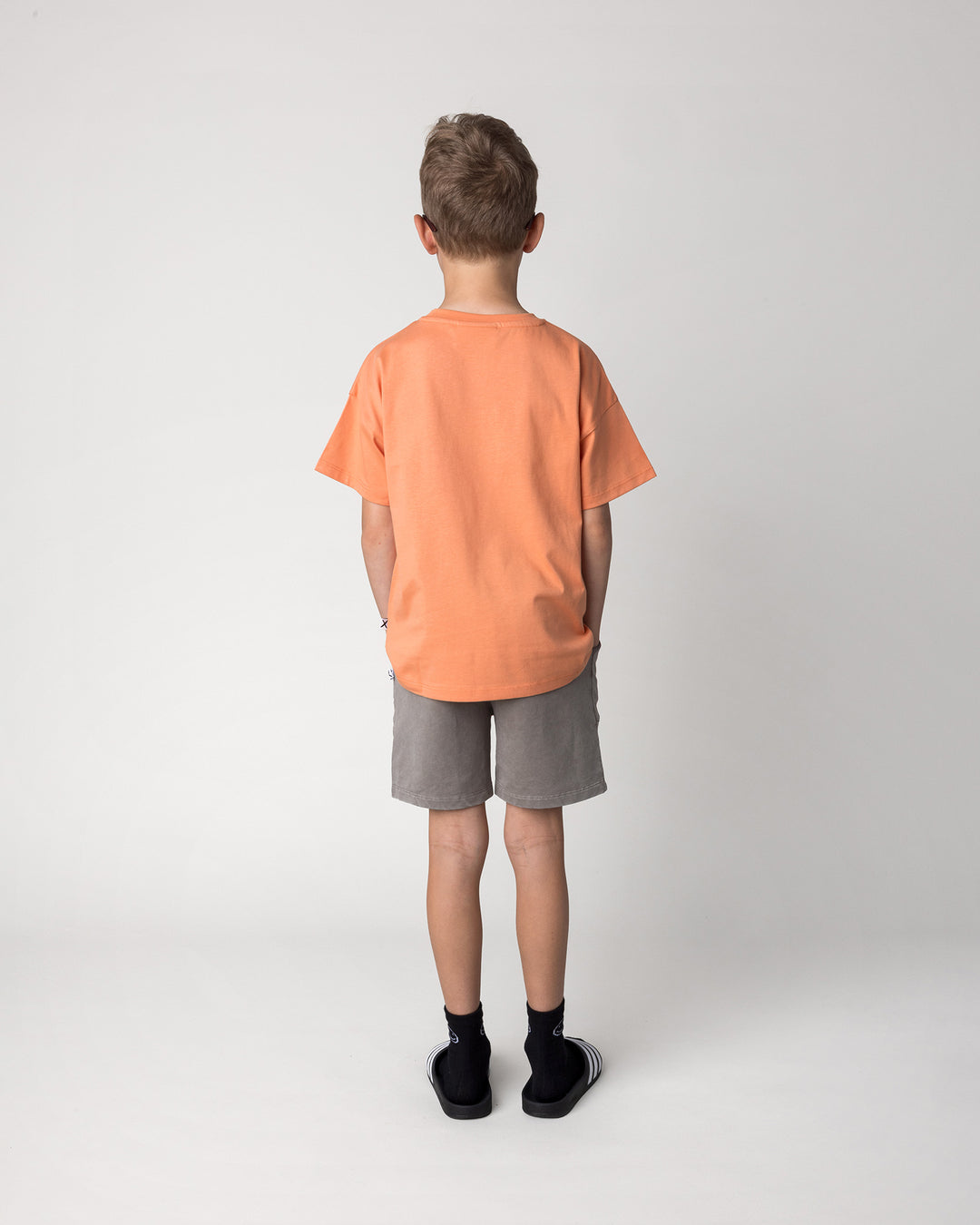 Kid wearing the Blasted Track Short in grey wash with the Vacay Crayfish Tee in light orange, back view