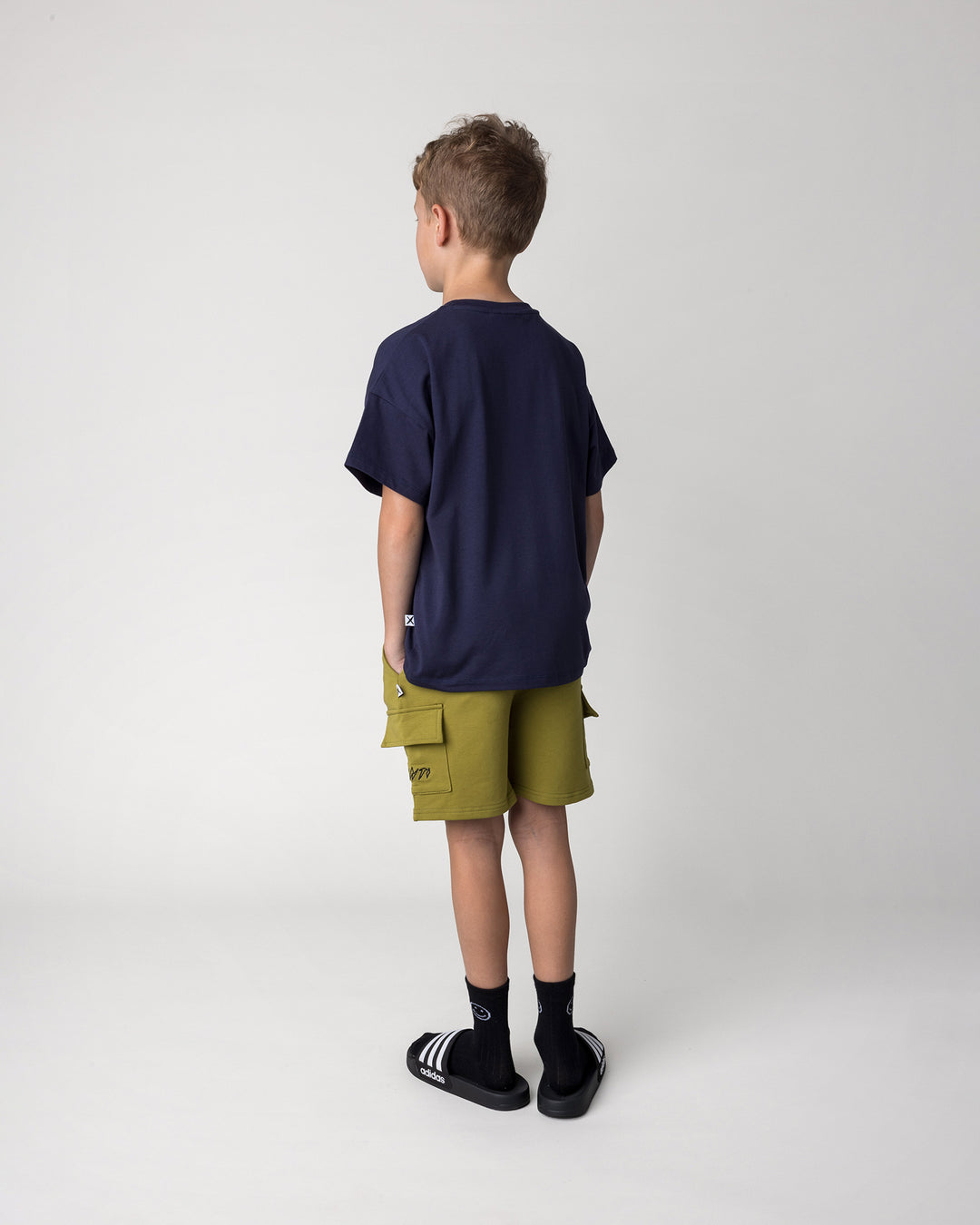 Kid wearing the Deluxe Cargo Short in olive with the Badges Tee in ink, back view