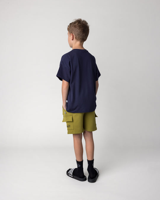 Kid wearing the Deluxe Cargo Short in olive with the Badges Tee in ink, back view