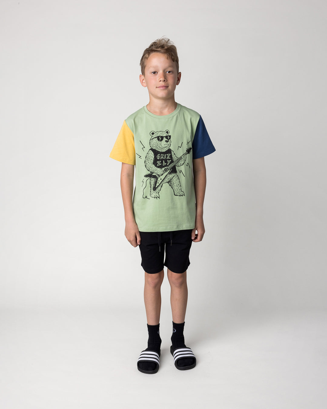 Kid wearing the Hero Short in black with the Grizzly Tee in sage, yellow and midnight, front view
