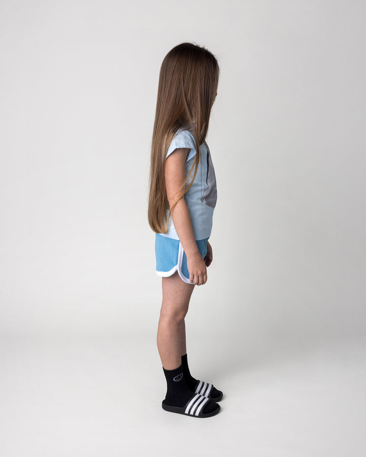 Kid wearing the Flower Penguin Tee in light blue with the Track Star Short in ocean, side view
