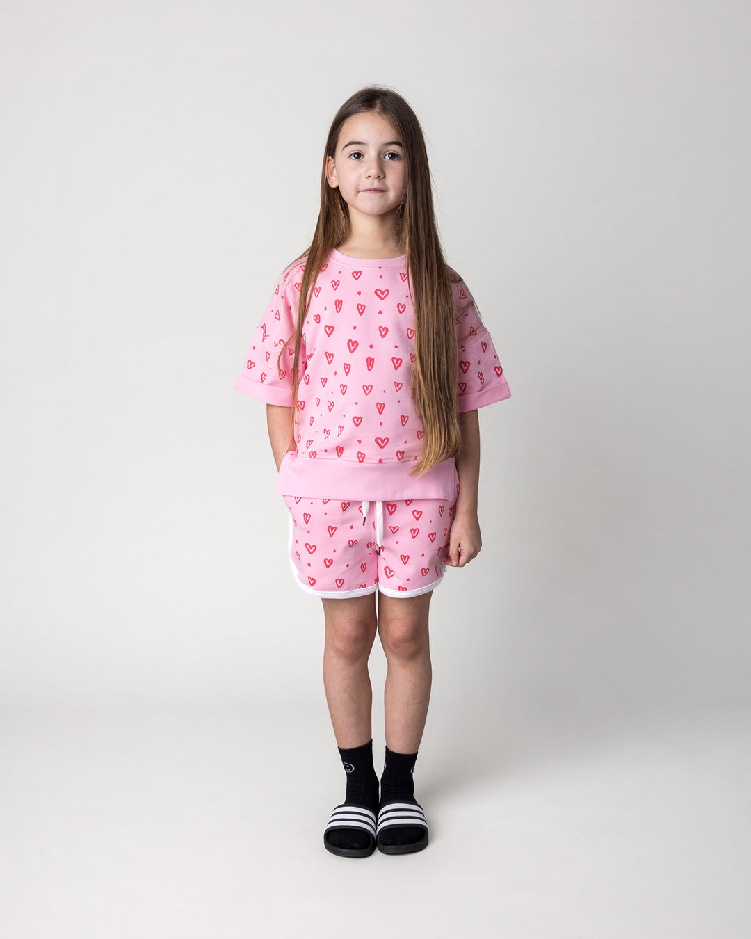 Kid wearing the Hearts Short Sleeve Crew in pink with the Hearts Short in pink and white, front view