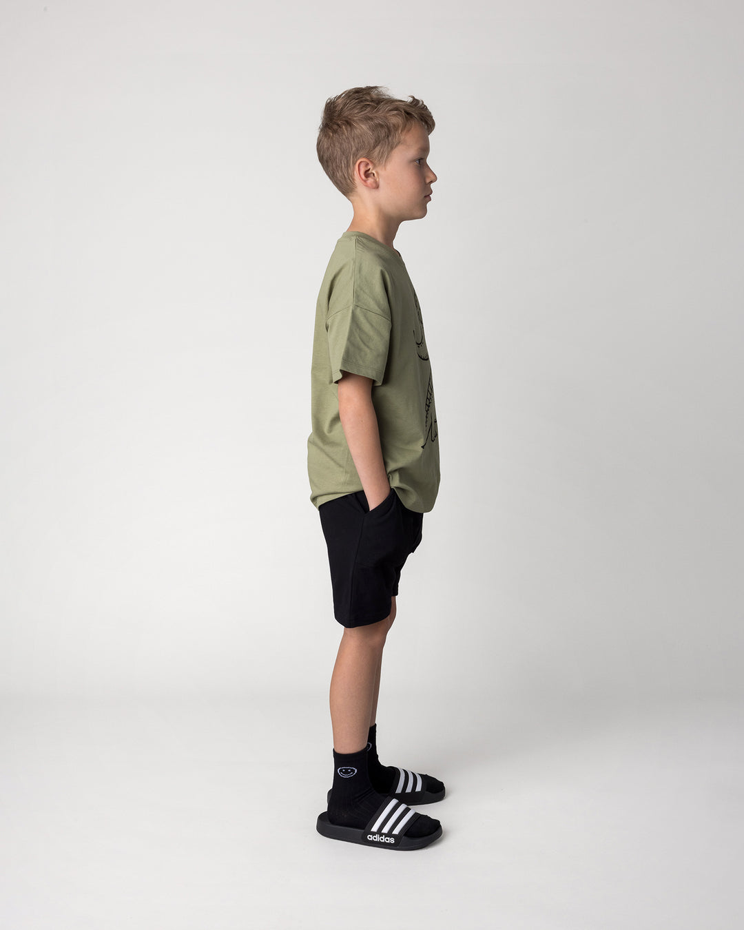 Kid wearing the Dino Ride Tee in light khaki with the Rib Pocket Short in black, side view
