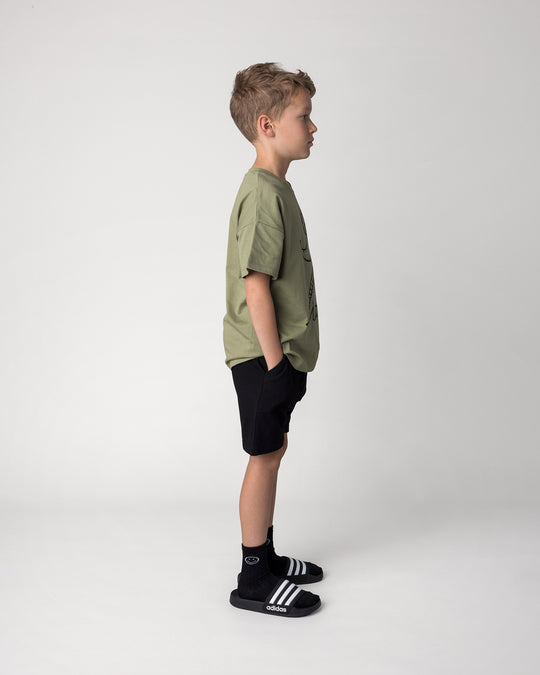 Kid wearing the Dino Ride Tee in light khaki with the Rib Pocket Short in black, side view