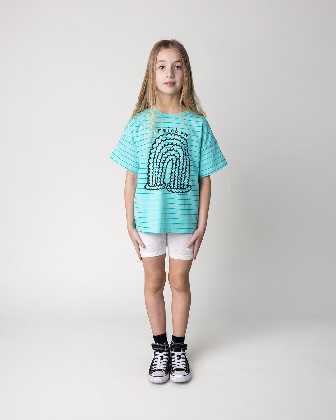 Kid wearing the Happy Rainbow Tee in teal stripe with the Velvet Bike Pant in cream, front view