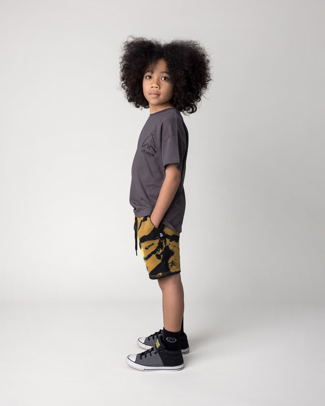 Kid wearing the Just Hanging Tee in dark grey with the Duo Tie Dye Short in lemon and rust, side view