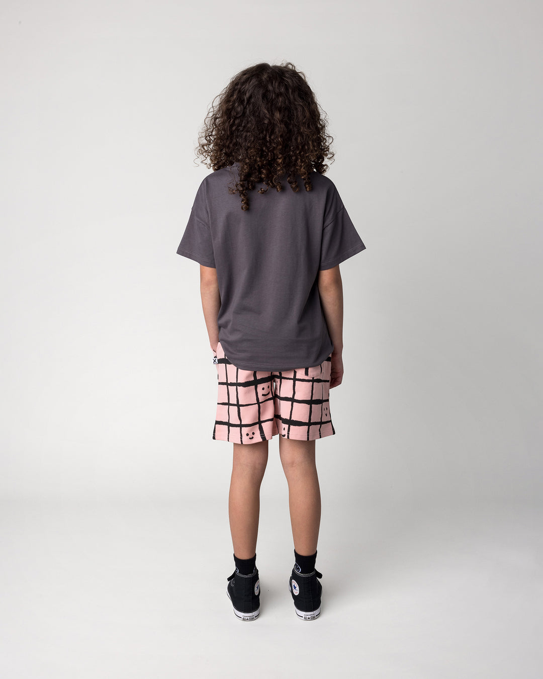 Kid wearing the Just Hanging Tee in dark grey with the Happy Squares Short in dusty pink, back view
