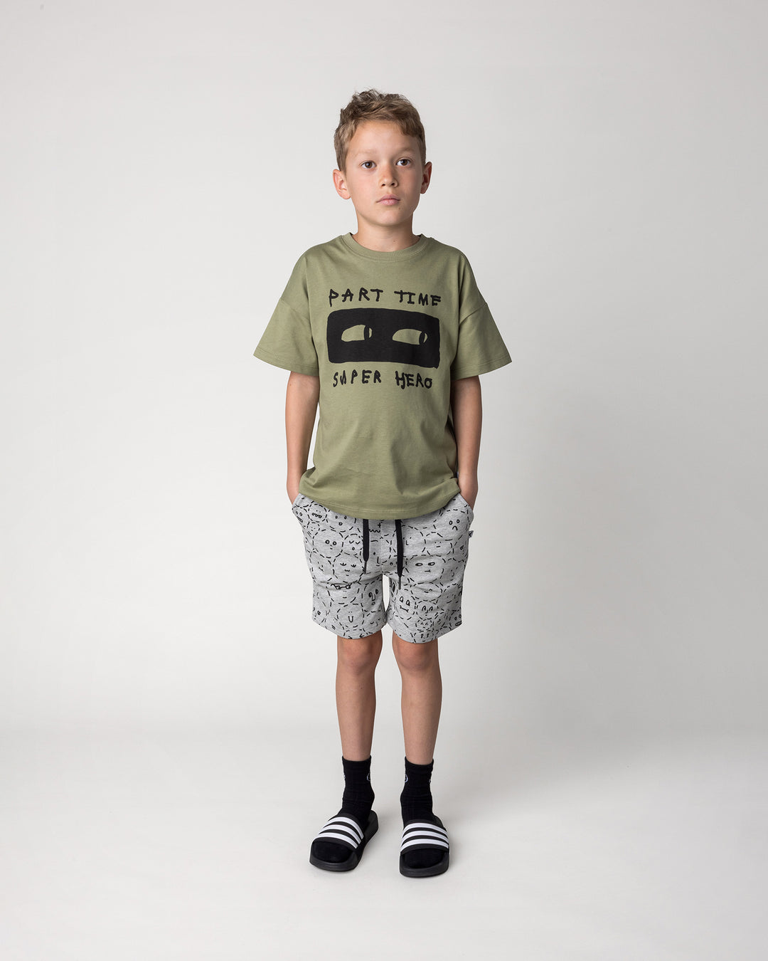 Kid wearing the Part Time Super Hero Tee in light khaki with the Different Faces Short in grey marle and black, front view