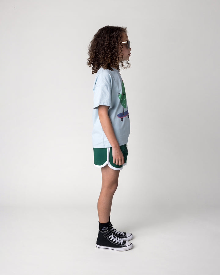 Kid wearing the Skate Cactus Tee in light blue with the Sprint Short in kelly green and white, side view