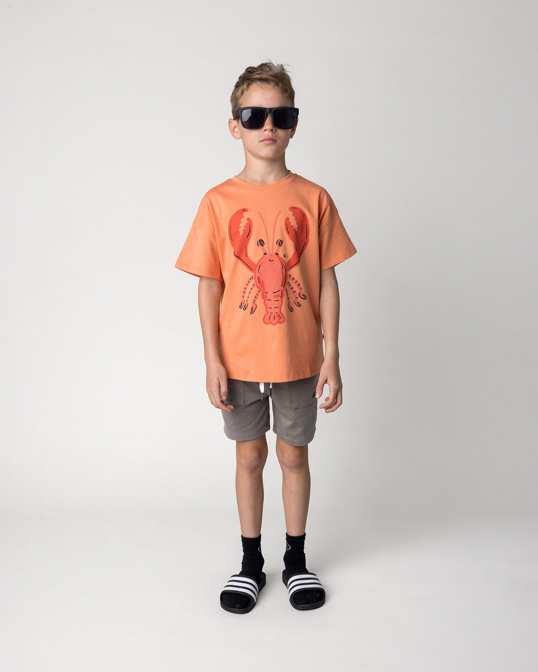 Kid wearing the Vacay Crayfish Tee in light orange with the Blasted Track Short in grey wash, front view