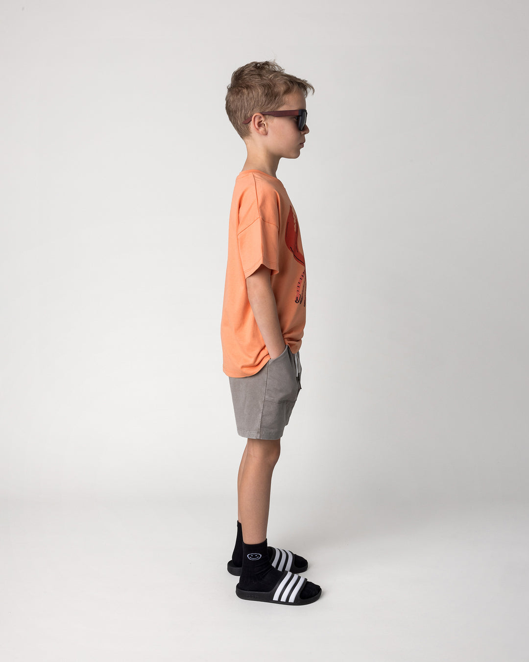 Kid wearing the Vacay Crayfish Tee in light orange with the Blasted Track Short in grey wash, side view