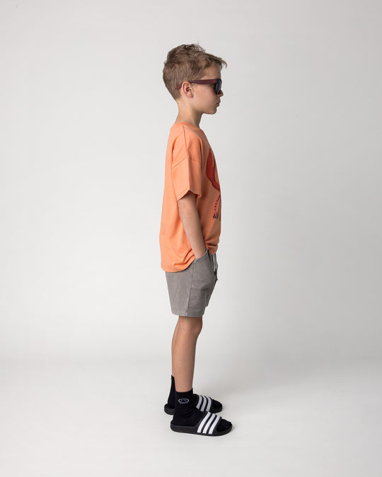 Kid wearing the Vacay Crayfish Tee in light orange with the Blasted Track Short in grey wash, side view