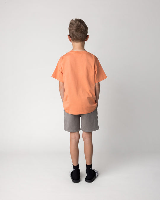 Kid wearing the Vacay Crayfish Tee in light orange with the Blasted Track Short in grey wash, back view