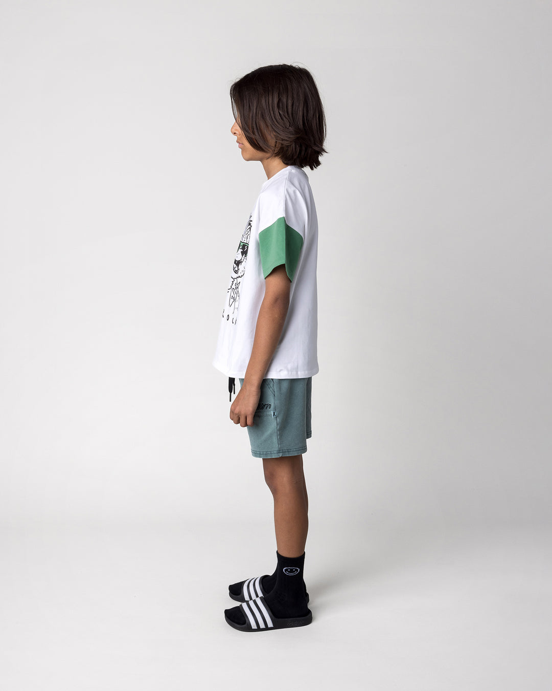 Kid wearing the Werewolf Rockstar Tee in off white, mustard and turf with the Blasted Track Short in forest wash, side view