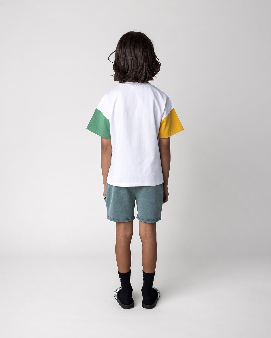 Kid wearing the Werewolf Rockstar Tee in off white, mustard and turf with the Blasted Track Short in forest wash, back view