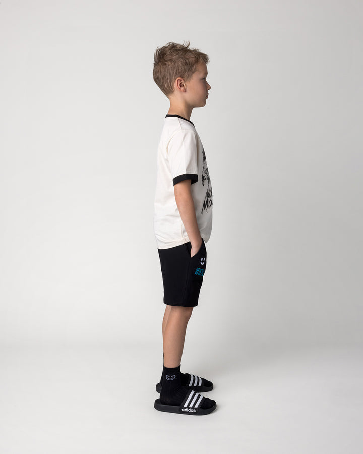 Kid wearing the Mo-Hawk Tee in cream and black with the Badges Short in black, side view