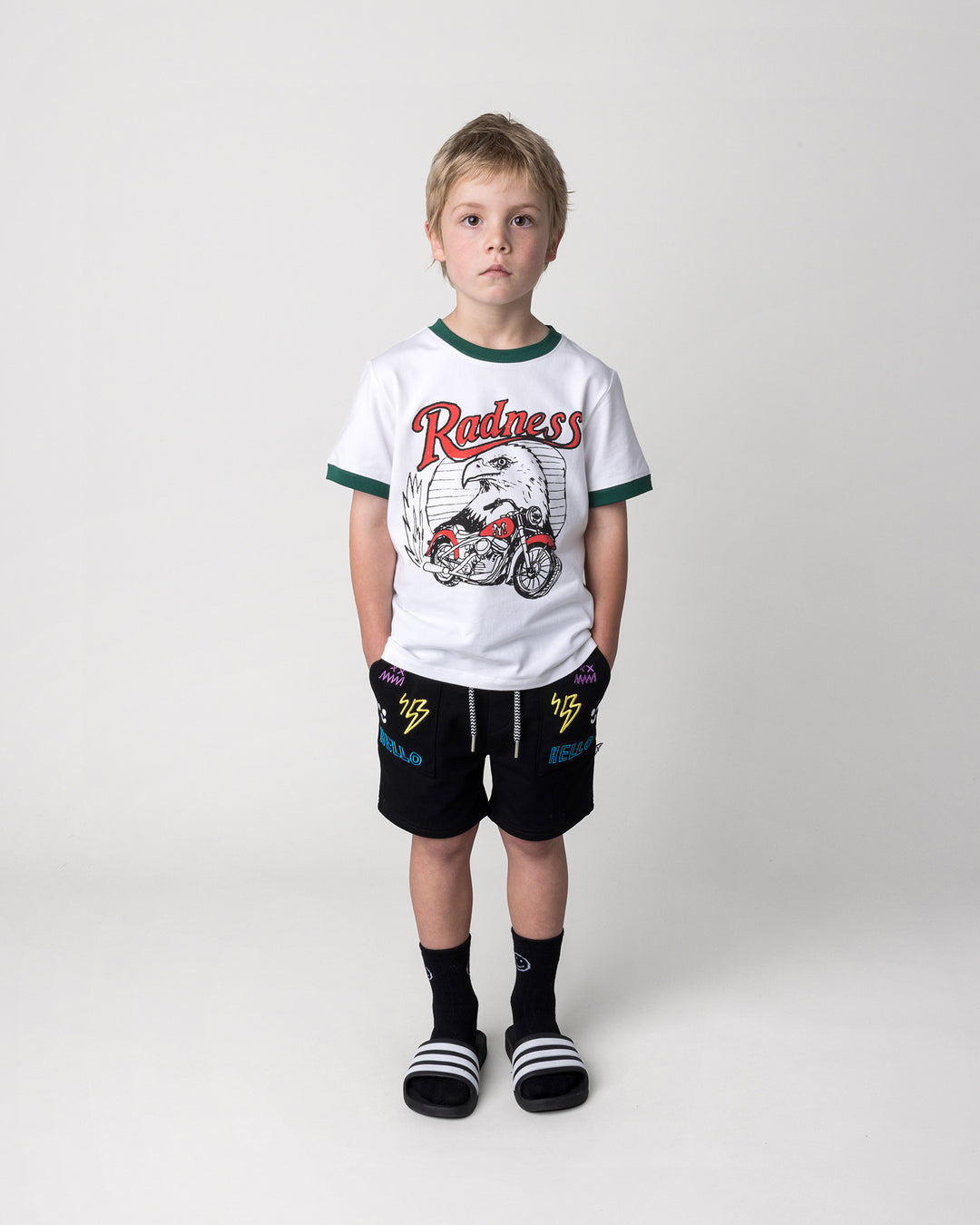Kid wearing the Radness Tee in white and jungle with the Badges Short in black, front view