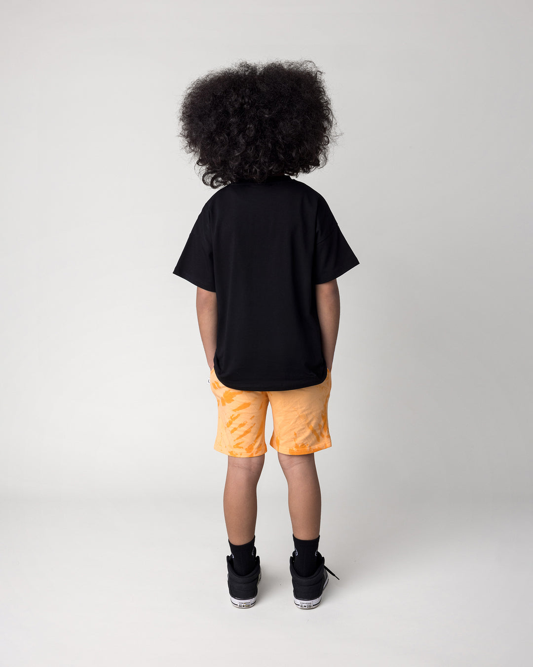 Kid wearing the Badges Tee in black with the Splatter Short in orange, back view
