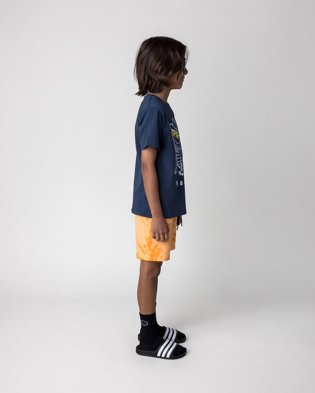 Kid wearing the Howdy Lizard Tee in ink with the Splatter Short in orange, side view
