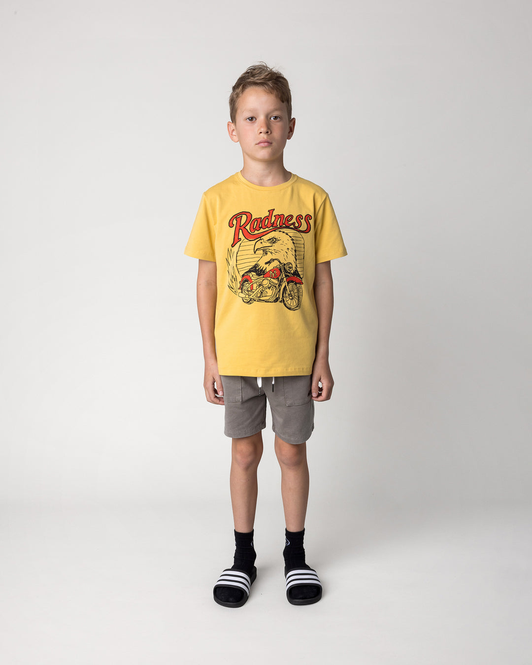 Kid wearing the Radness Tee in dull yellow with the Blasted Track Short in grey wash, front view