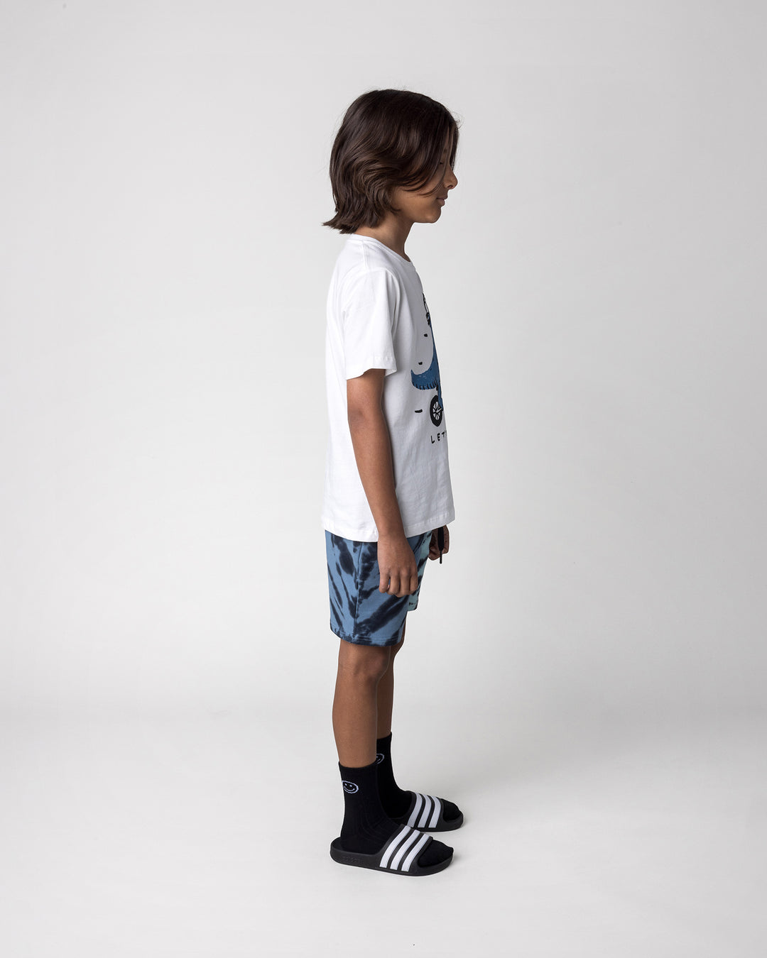 Kid wearing the Rex Rider Tee in off white with the Duo Tie Dye Short in blue and aqua, side view