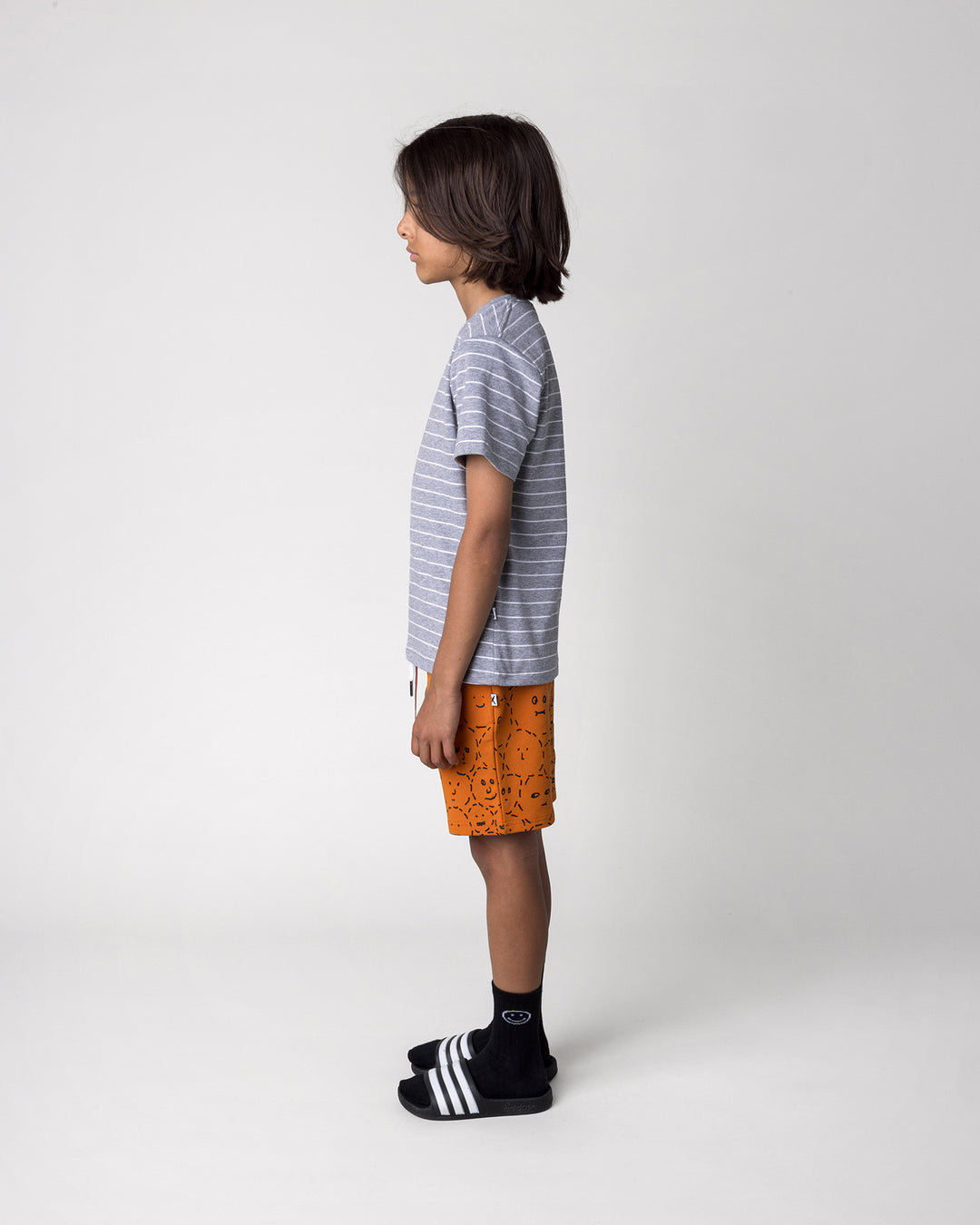 Kid wearing the Super Cool Tee in grey stripe with the Different Faces Short in burnt orange, side view