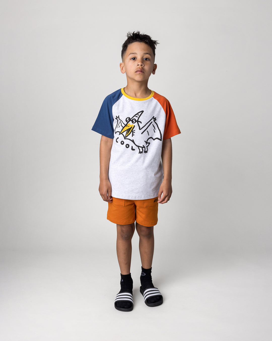 Kid wearing the Cool Pterodactyl Tee in white marle and multi with the Track Short in burnt orange, front view