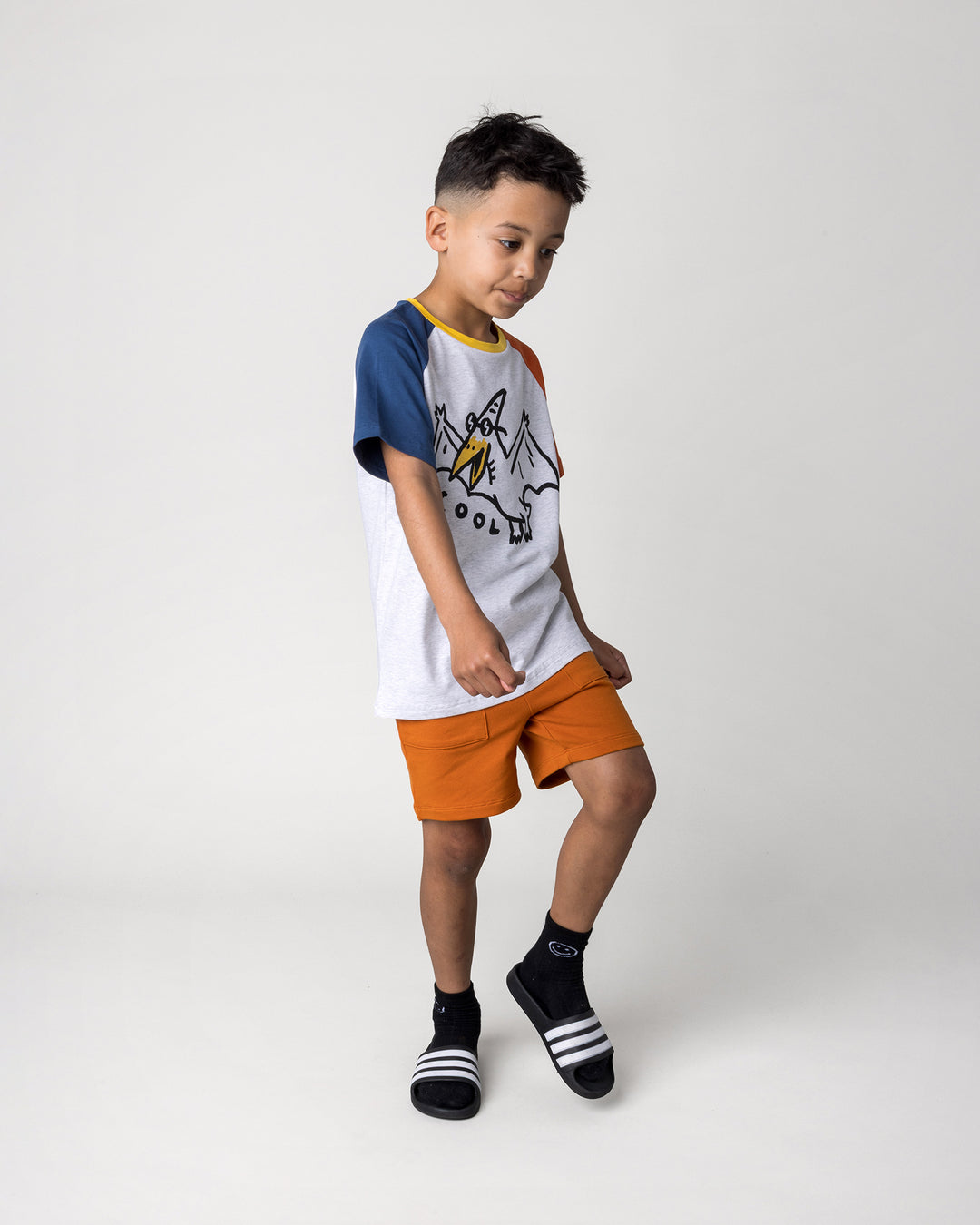 Kid wearing the Cool Pterodactyl Tee in white marle and multi with the Track Short in burnt orange, side view