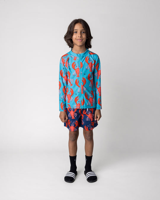 Kid wearing the Vacay Crayfish Rashie in teal with the Vacay Crayfish Boardies in dark blue, front view