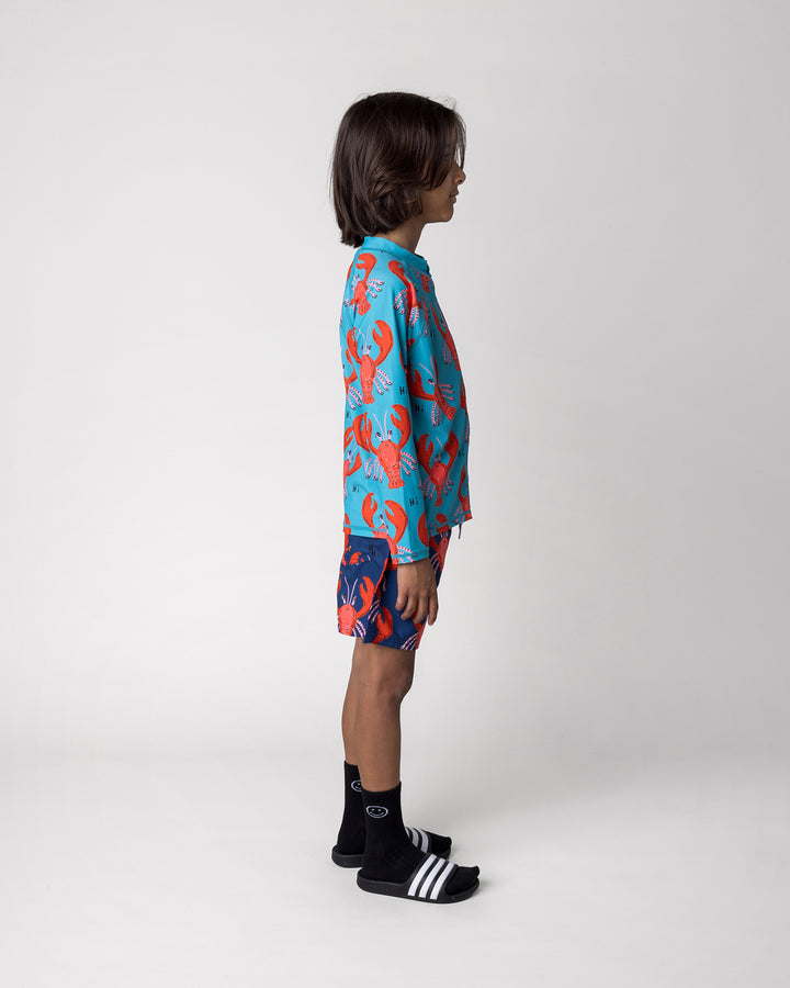 Kid wearing the Vacay Crayfish Rashie in teal with the Vacay Crayfish Boardies in dark blue, side view