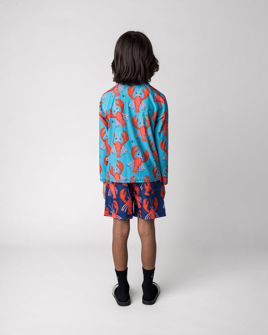 Kid wearing the Vacay Crayfish Rashie in teal with the Vacay Crayfish Boardies in dark blue, back view